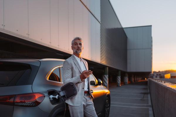 A businessman holding smartphone while charging car at electric vehicle charging station, closeup.