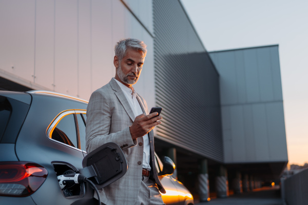 A businessman holding smartphone while charging car at electric vehicle charging station, closeup.