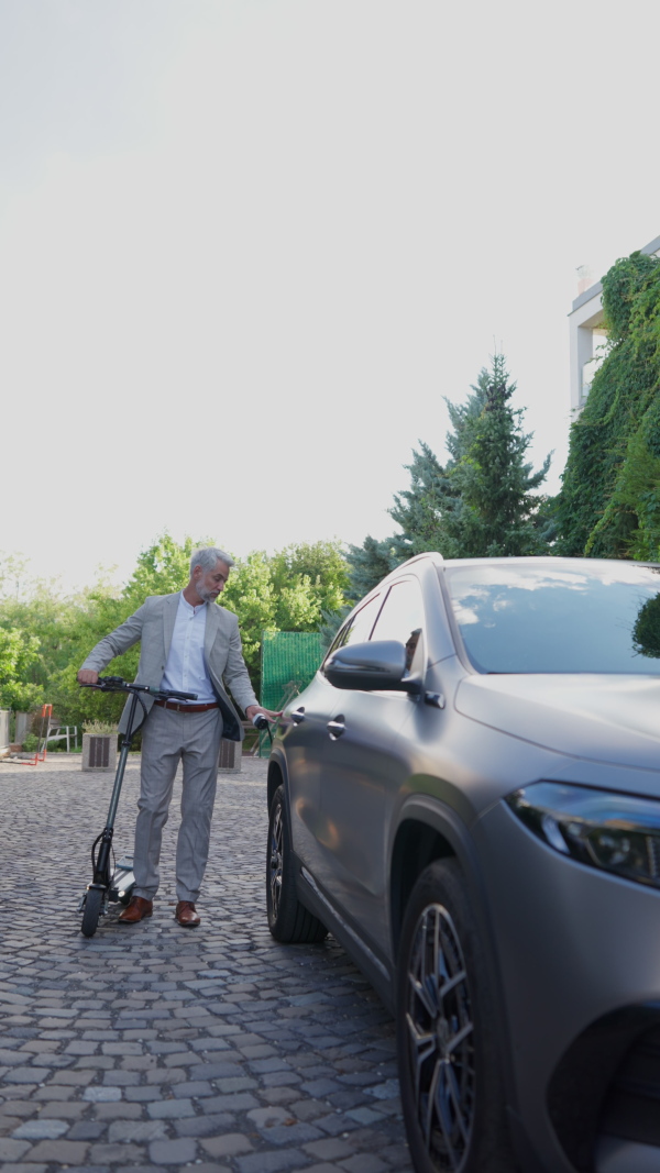 Mature businessman with electric scooter charging electric car in driveway. Vertical shot.