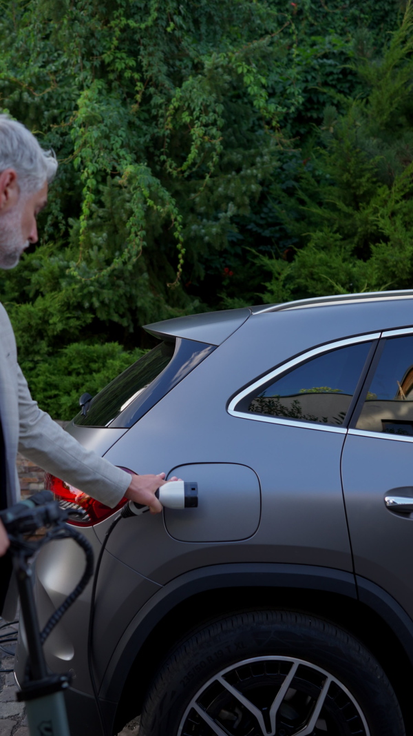 Mature man charging battery of electric car in driveway. Vertical shot.