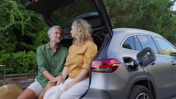 Happy mature man and woman sitting in car trunk while charging electric car.