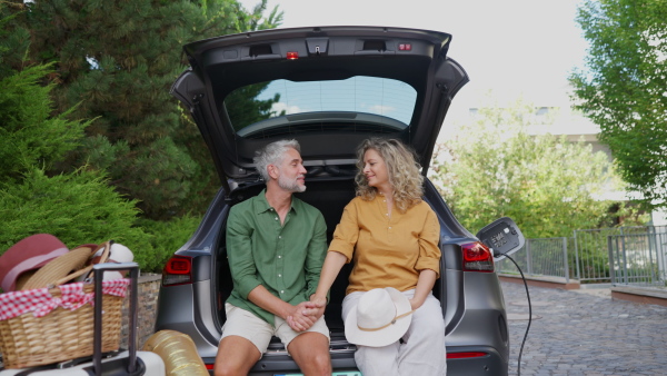 Portrait of mature couple sitting together in open car trunk, while charging battery at electric car before going on holiday trip. Suitcase in foreground. Looking into camera.