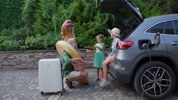 Family charging electric car, preparing for holiday travel in driveway, packing suitcases into trunk.