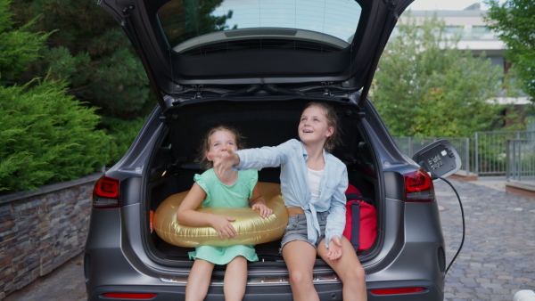 Two happy girls sititng in open electric car trunk, preparing for holiday travel while charging car.