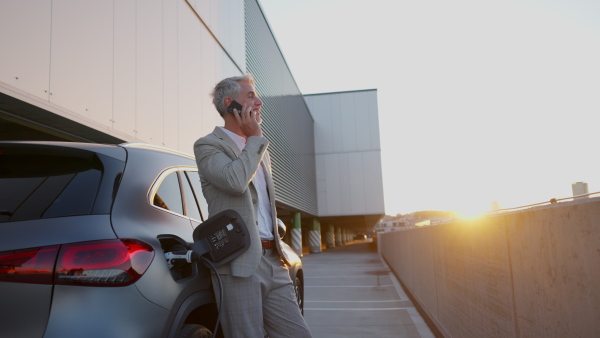 Businessman charging battery of electric car and using mobile phone on rooftop of modern building with sunset in background.