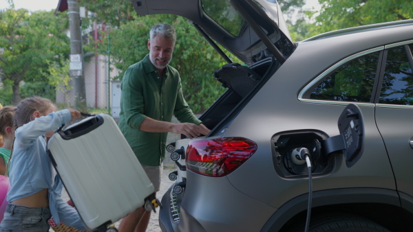 Family charging electric car, preparing for holiday travel, putting suitcases in car trunk.