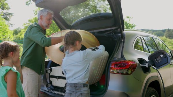 Father with kids packing suitcases an swimming gear into electric car trunk while charging battery before travel.