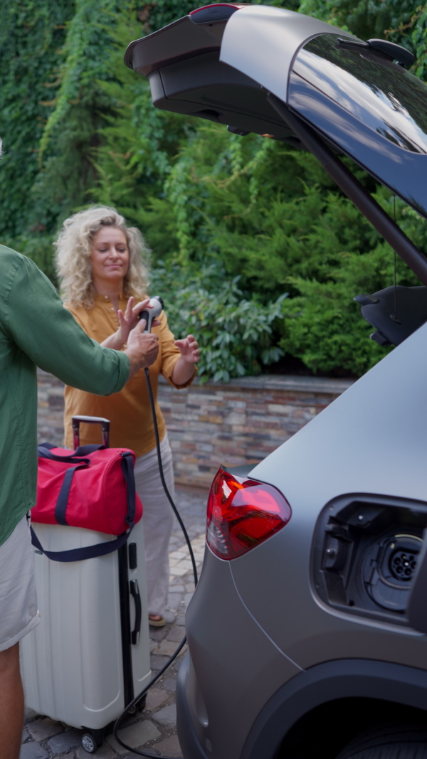 Woman passing charger of electric car to man, charging electric car in frontyard before travelling.