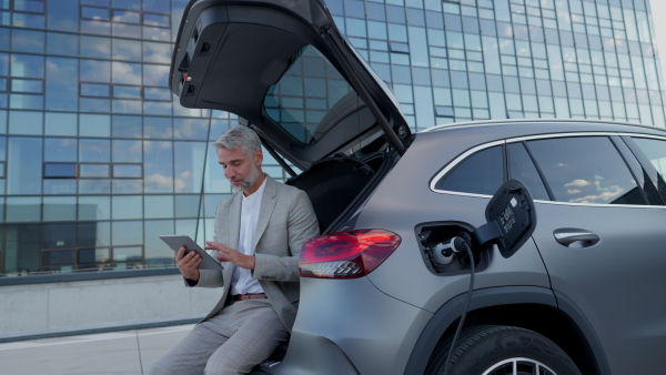 Portrait of mature businessman using digital tablet while sitting in car, charging electric car battery in front of modern office building.