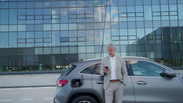 Mature businessman in suit charging battery of electric car while using mobile phone in front of modern office building.