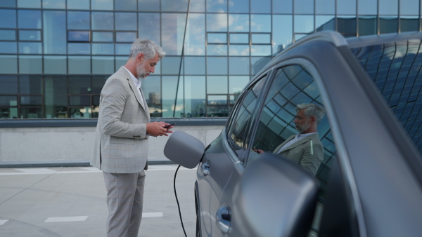 Man charging electric car in front of office building while messaging on mobile phone.