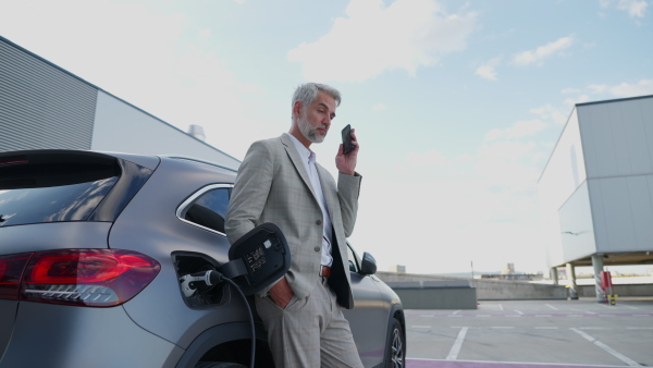 Mature businessman charging battery of electric car using mobile phone on rooftop.