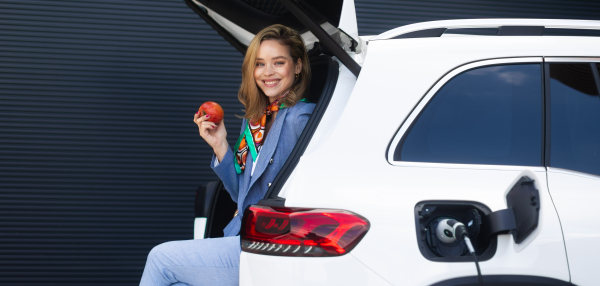 Young woman waiting for her electric car charging, sitting in a car trunk and eating an apple.