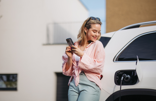 Young woman with smartphone charging electric car in home, sustainable and economic transportation concept.