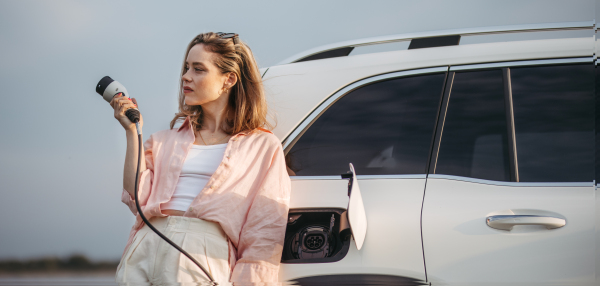 Young woman holding power supply cable from her car, prepared for charging it, sustainable and economic transportation concept.