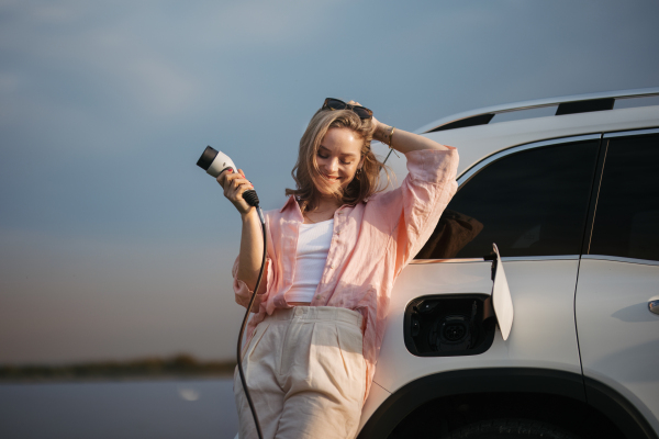 Young woman holding power supply cable from her car, prepared for charging it, sustainable and economic transportation concept.