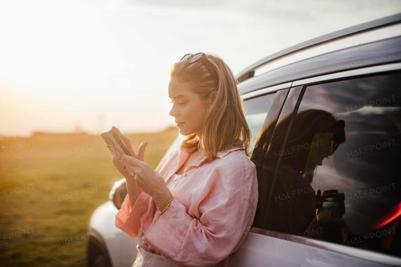 Young woman scrolling phone, leaning on her car in the nature,