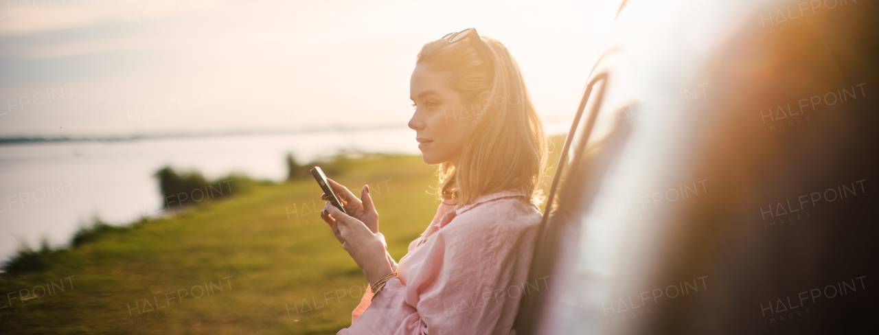 Young woman scrolling phone, leaning on her car in the nature,