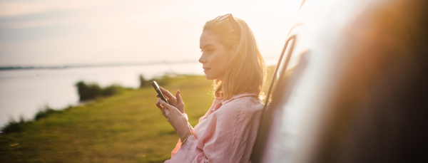 Young woman scrolling phone, leaning on her car in the nature,