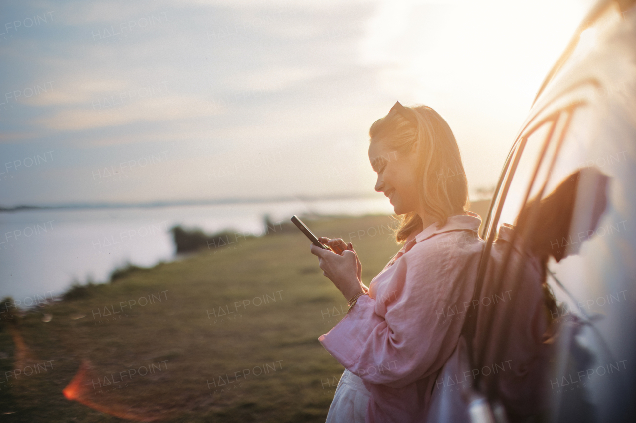 Young woman scrolling phone, leaning on her car in the nature,