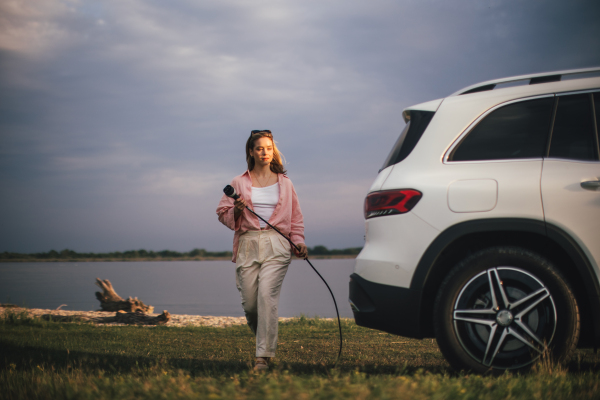 Young woman holding power supply cable from her car, prepared for charging it, sustainable and economic transportation concept.