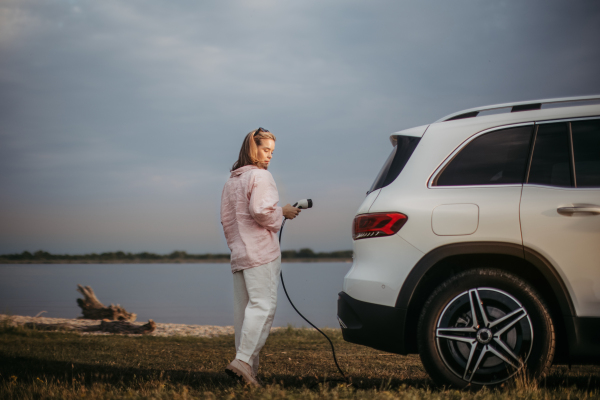 Young woman holding power supply cable from her car, prepared for charging it, sustainable and economic transportation concept.