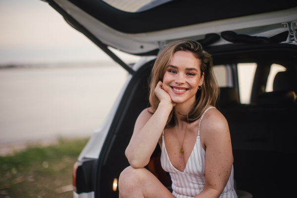 Young woman sitting in car trunk near lake, enjoying summer time.