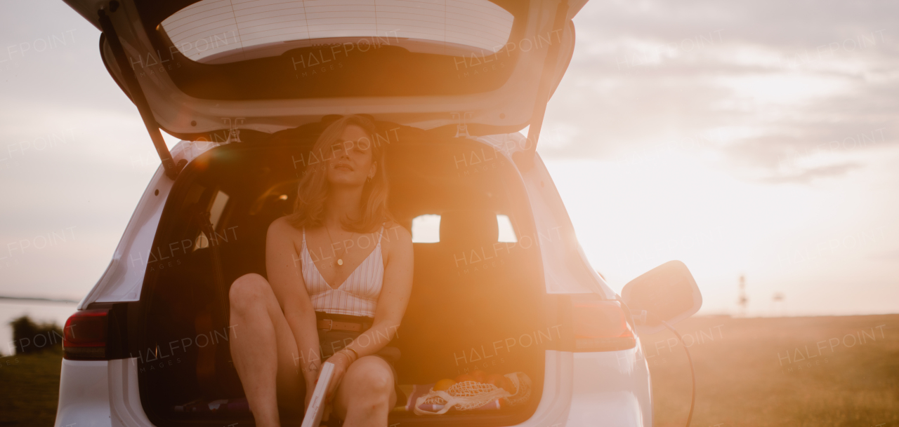 Young woman sitting in a car trunk, waiting for charging it.