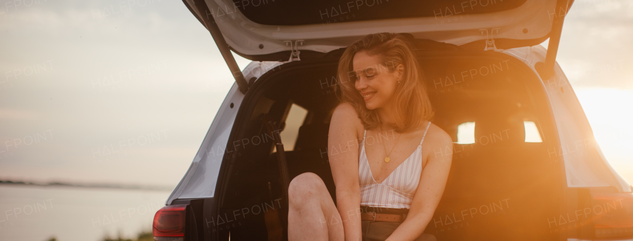 Young woman sitting in car trunk near lake, enjoying summer time.