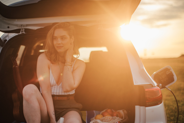 Young woman sitting in a car trunk, waiting for charging it.