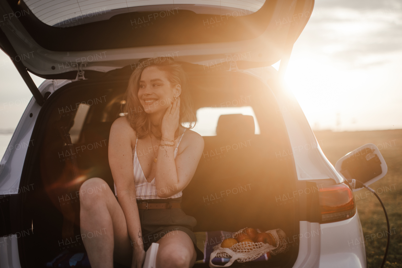 Young woman sitting in a car trunk, waiting for charging it.