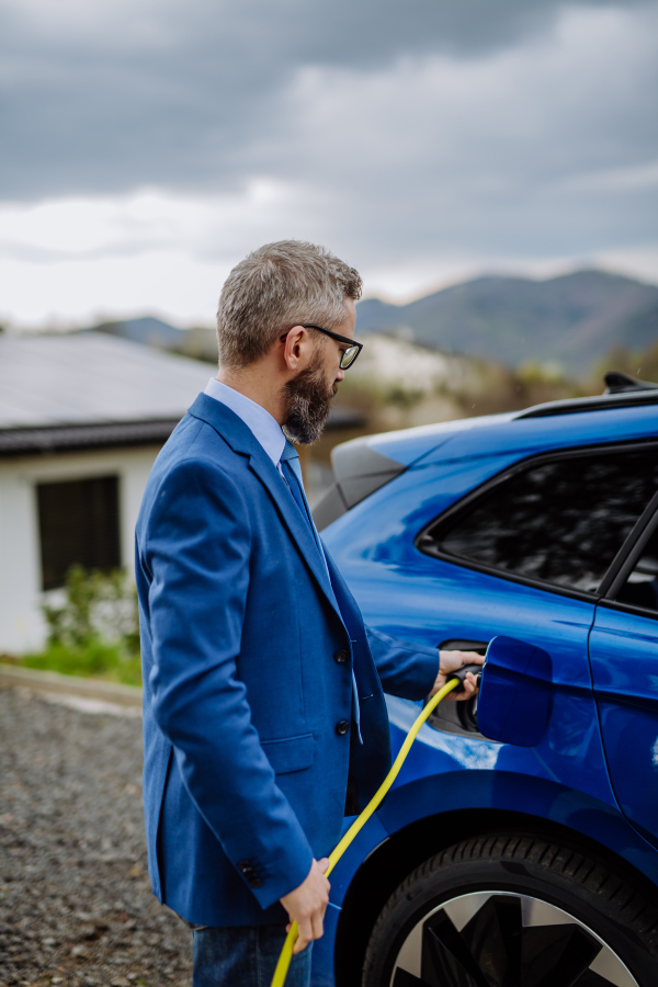 Mature businessman holding power supply cable and charging his new electric car.