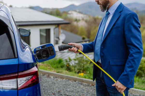 Mature businessman holding power supply cable and charging his new electric car.