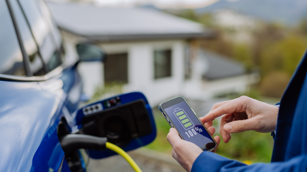 Close up of businessman holding phone, checking charging of his electric car.