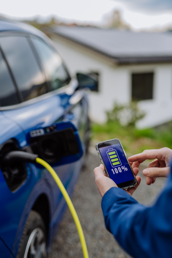 Close up of businessman holding phone, checking charging of his electric car.