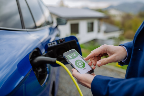 Close up of businessman holding phone, checking charging of his electric car.