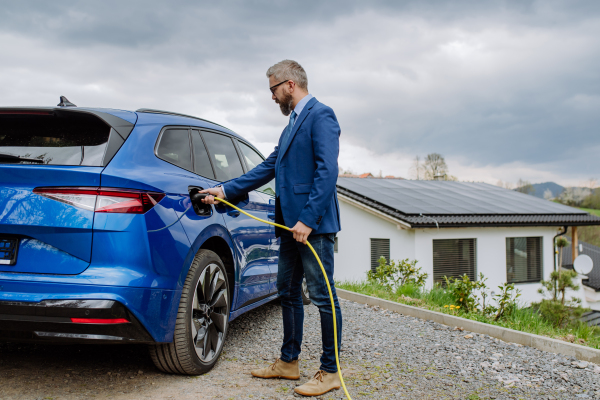 Mature businessman holding power supply cable and charging his new electric car.