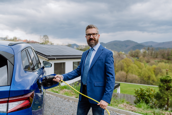 Mature businessman holding power supply cable and charging his new electric car.