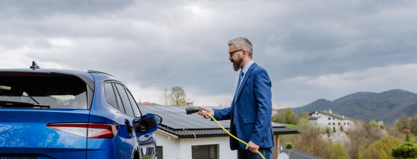 Mature businessman holding power supply cable and charging his new electric car.