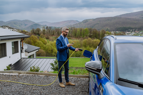 Mature businessman holding power supply cable, scrolling his phone and charging his new electric car.