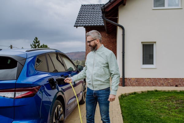 Mature man holding power supply cable and charging his new electric car.