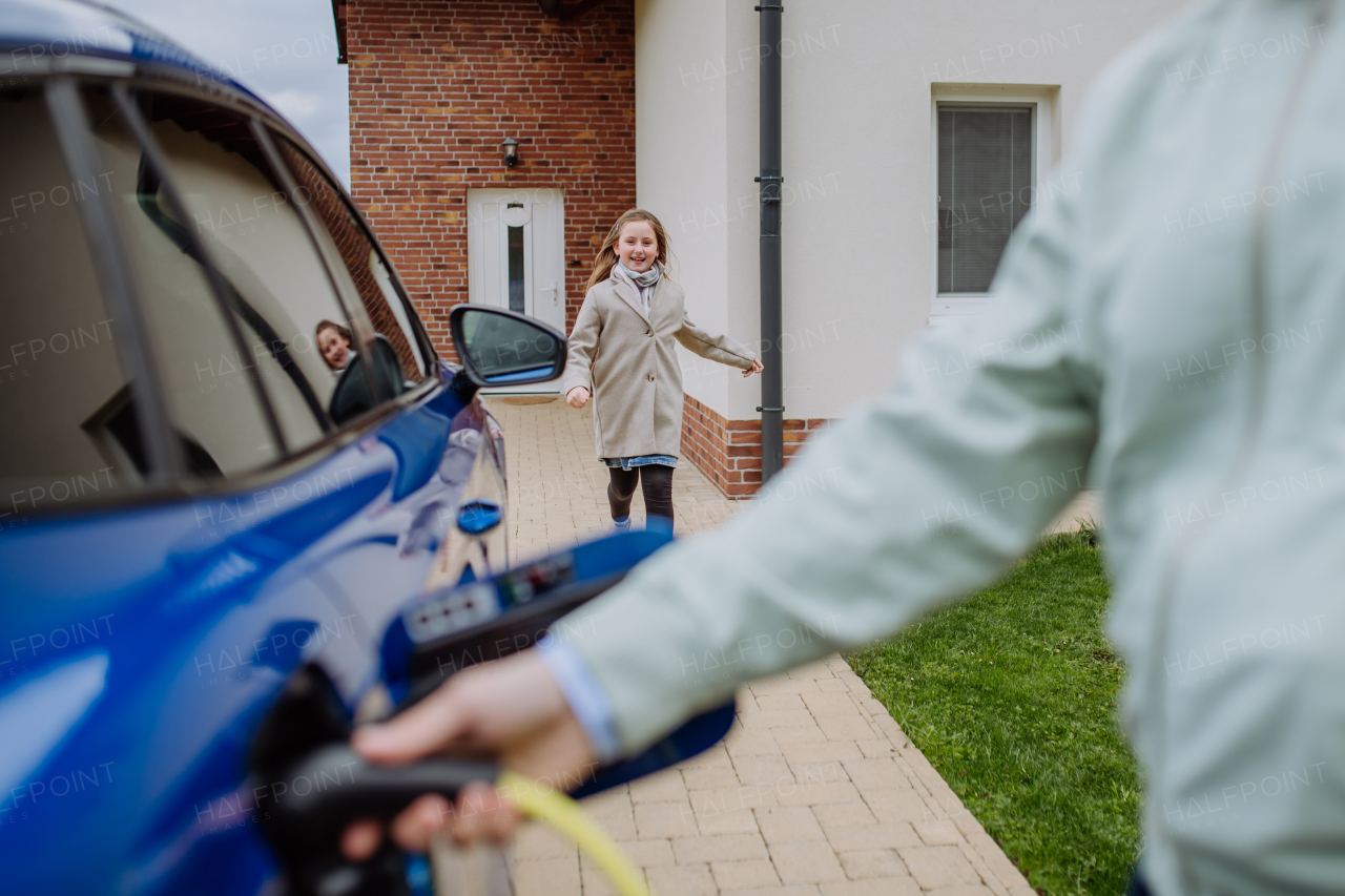 Close up of man holding power supply cable and charging his new electric car, his daughter running to him.