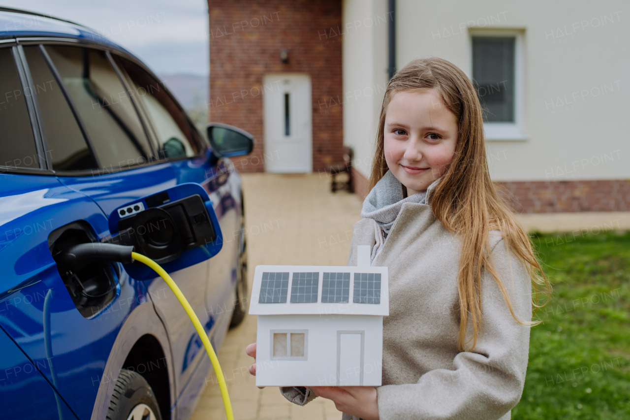 Little girl holding model of a paper house with solar panels, waiting for charging their electric car.