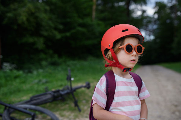 Portrait of little girl in red bicycle helmet waiting on forest path in park.