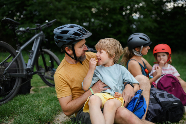 A young family with little children resting after bike ride, sitting on grass in park in summer.