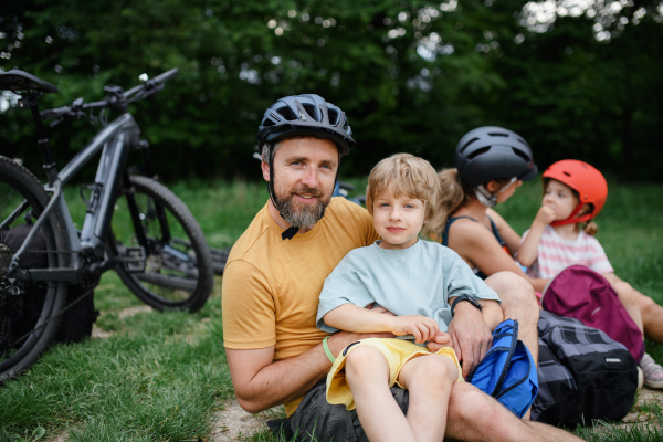 A young family with little children resting after bike ride, sitting on grass in park in summer.