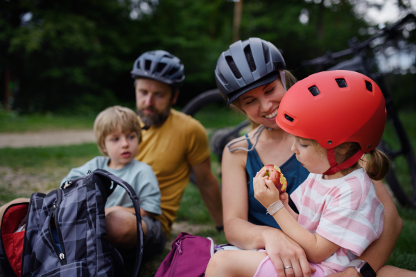 A young family with little children resting after bike ride, sitting on grass in park in summer.