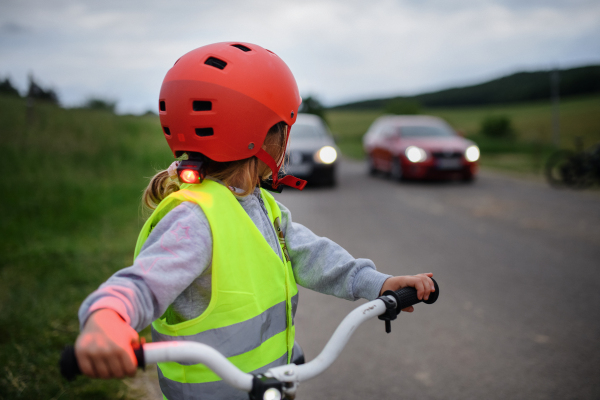 Rear view of little girl in reflective vest riding bike on road with cars behind her