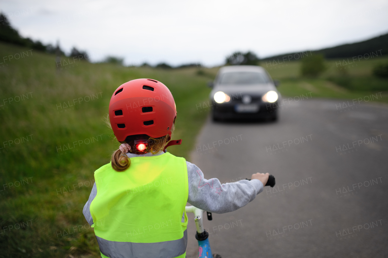 A rear view of child riding bike on road with car in front of her, road safety education concept.