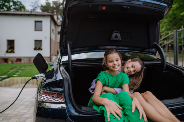 Happy sisters sitting in a car trunk and waiting for electric car charging.